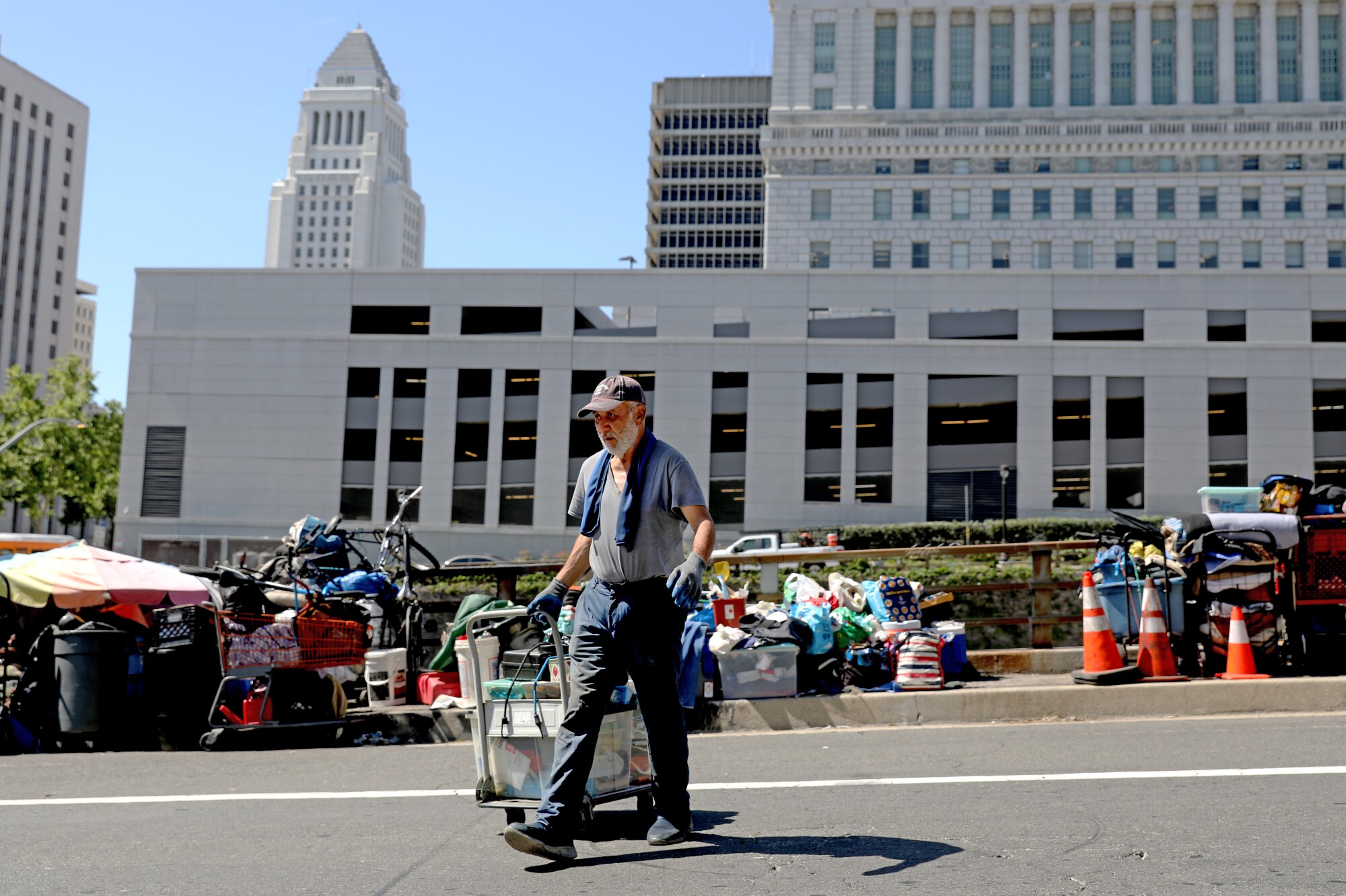 A man crosses the street pulling a handcart, with L.A. City Hall visible in the background.