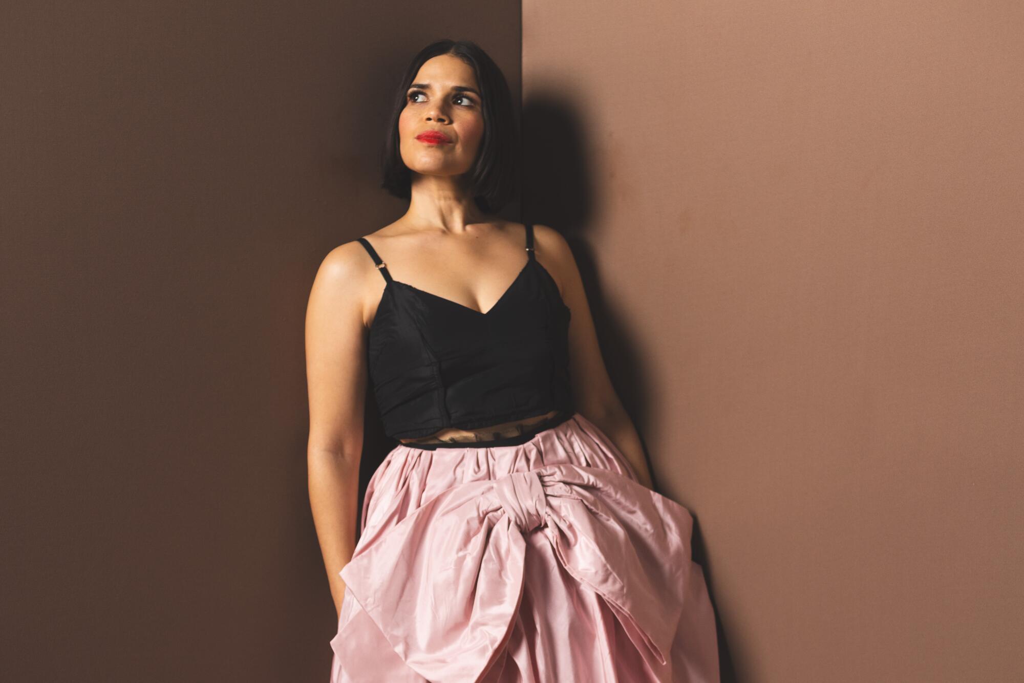 America Ferrera wears a black camisole top and pink skirt with a big bow, leaning against a wall and looking up.