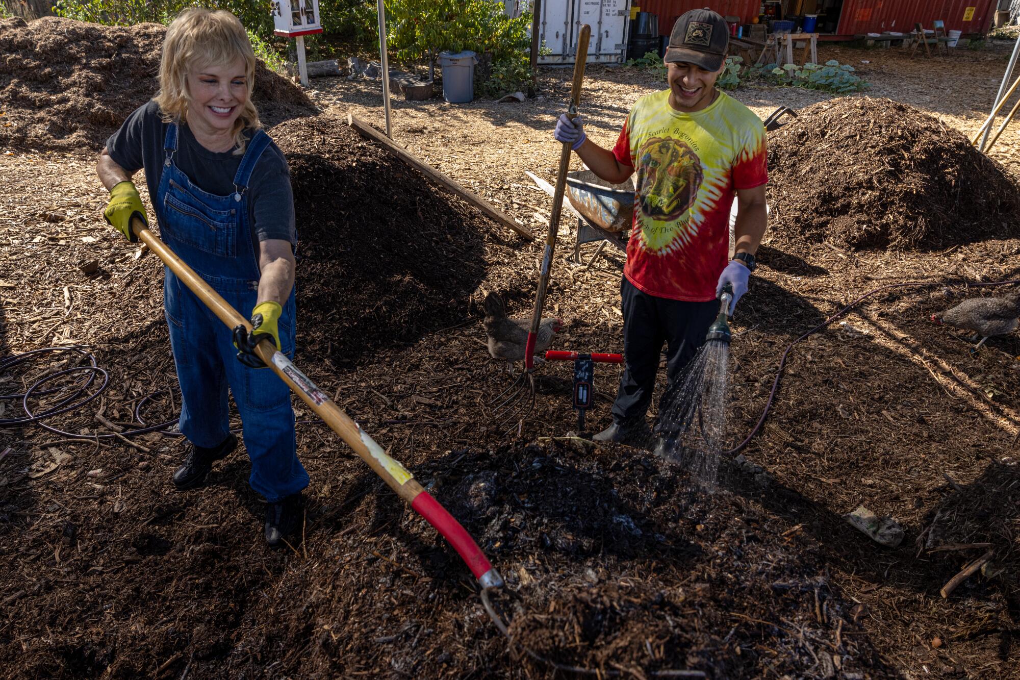 A person uses a garden forks to turn a pile of compost while another person waters the brown soil with a hose