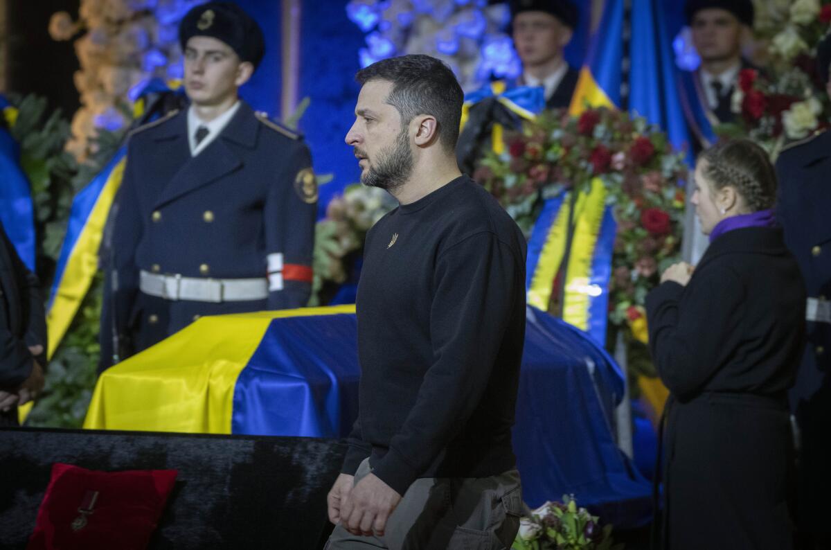 Ukrainian President Volodymyr Zelensky stands near men in uniform next to flag-draped coffins and flower wreaths.