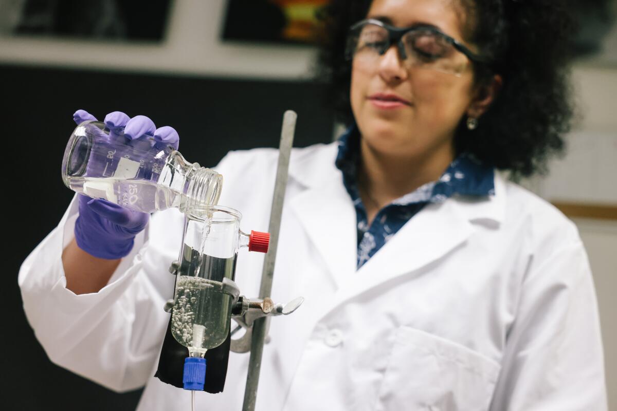 A woman in a lab coat and safety goggles pours liquid from one container to another in a lab setting.