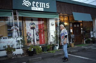 A man stands outside building whose front display shows baseball memorabilia. Its sign reads "Seems hair & spa."