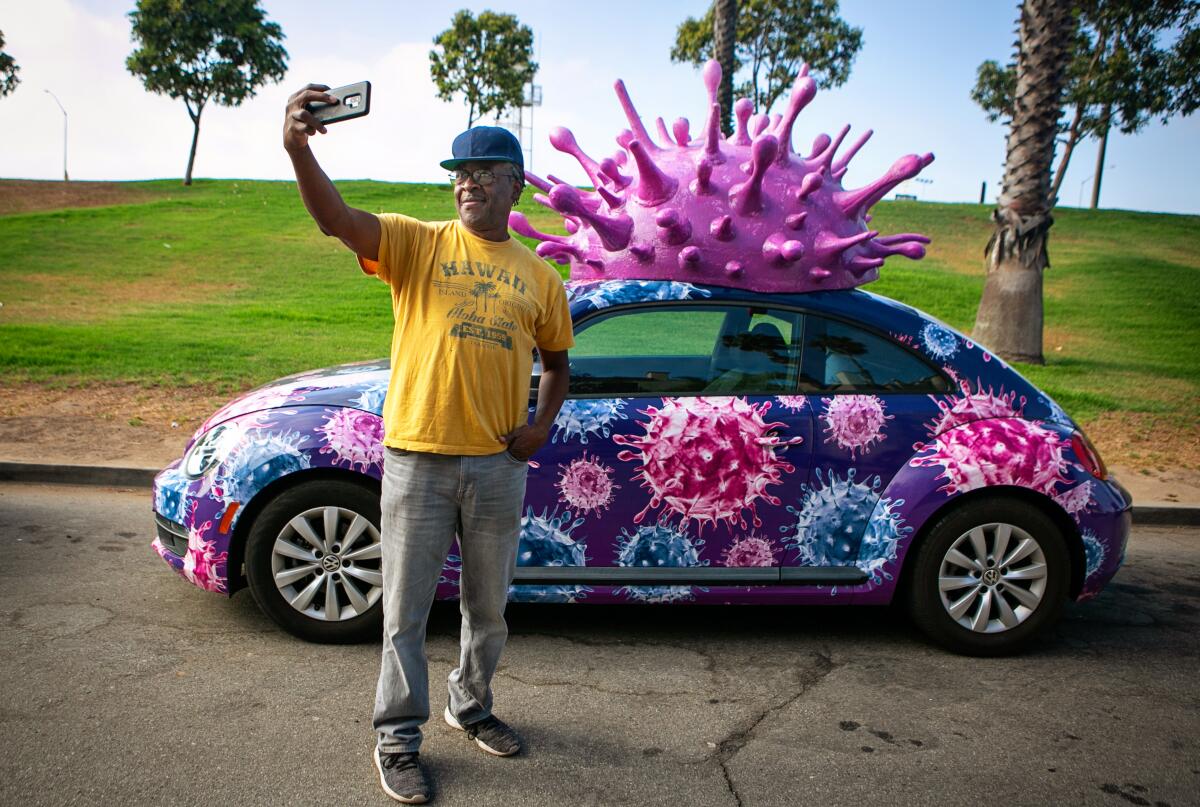 A man takes a selfie in front of a car decorated with a plastic coronavirus
