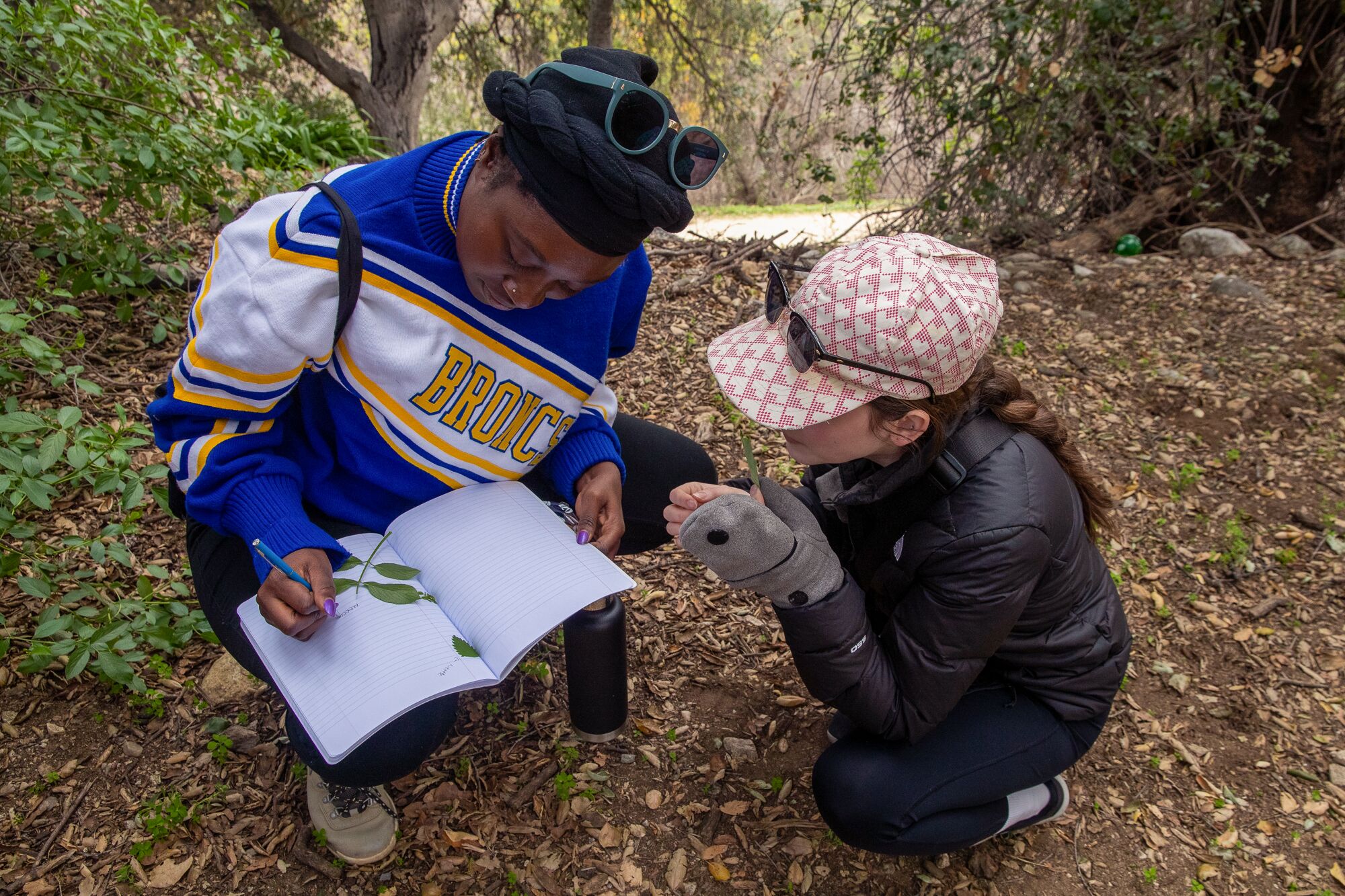 two women kneel down in a forest, the one on the left writing in a notebook 