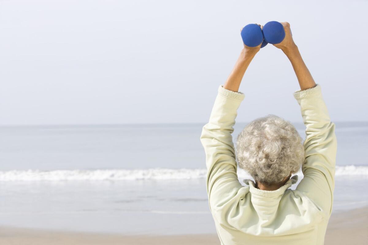 A woman with white hair pictured from behind on a beach, facing the water and holding two blue dumbbells up high