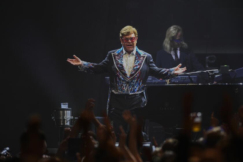 Elton John opens his arms onstage while performing in front of a piano