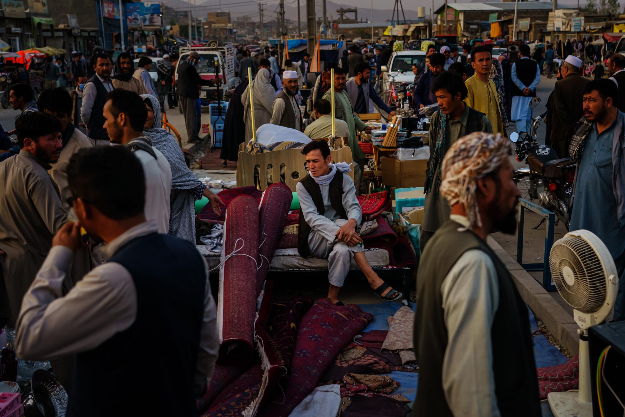 A person sits on a bed among people walking in a street bazaar.