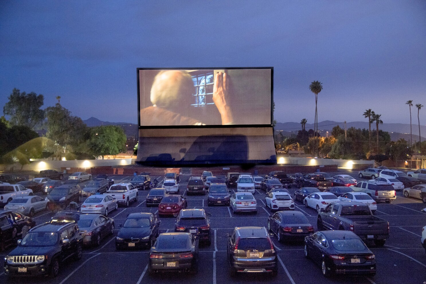 riverside drive thru movie theater with multiple cars parked in the drive thru lot staggered a large movie screen with movie playing with trees mountain and the city in the background and lights