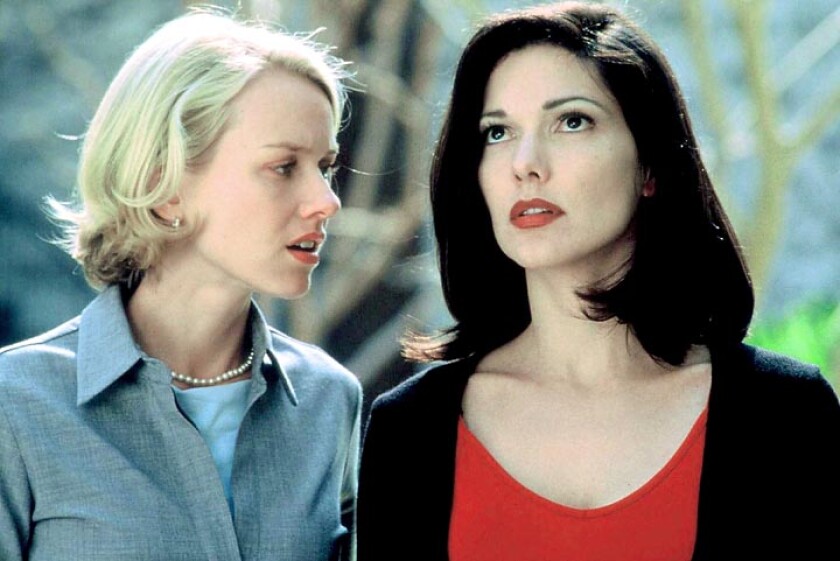 A blonde woman on the left wears a blue blouse and a pearl necklace and a brunette woman on the right wears a red blouse.