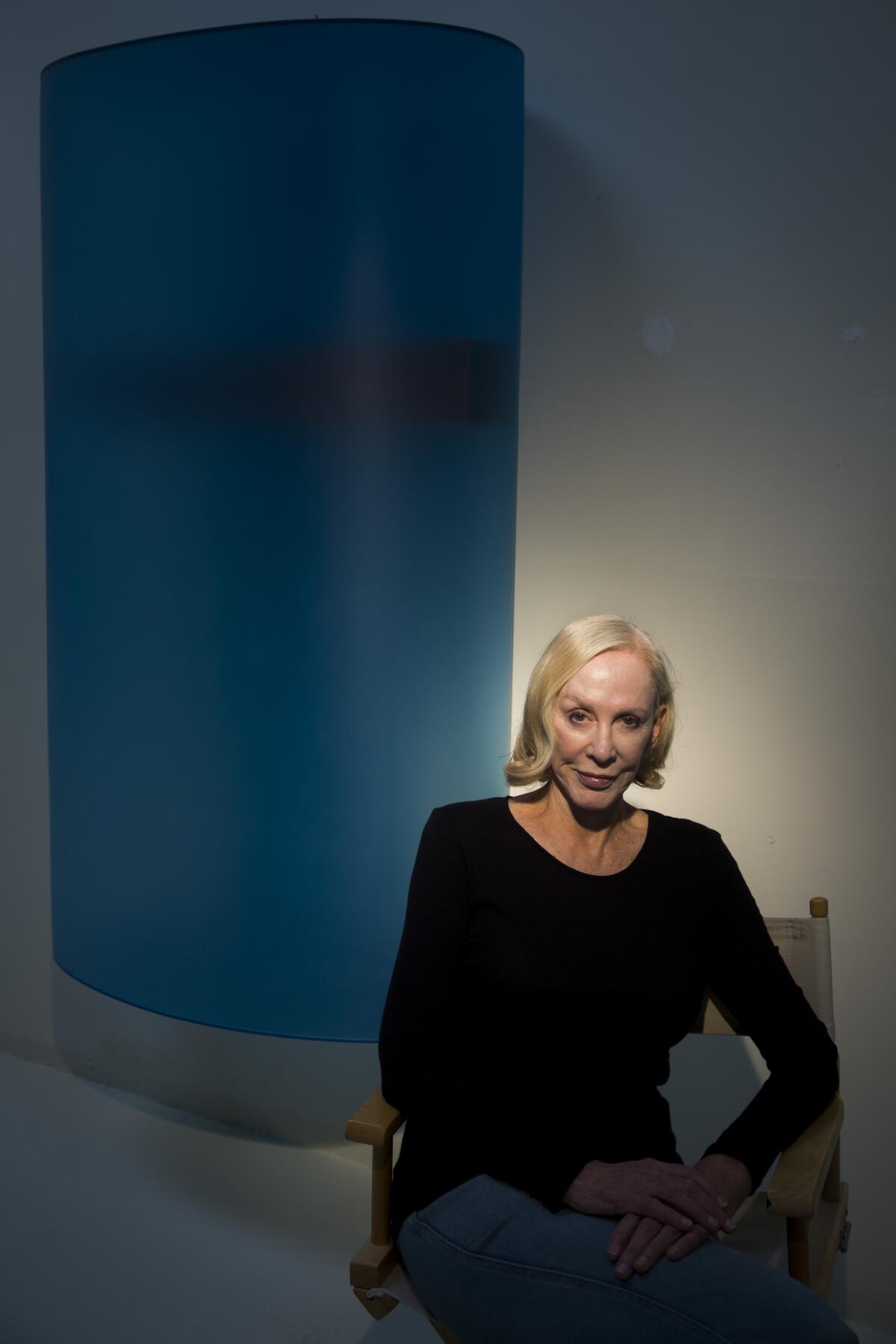 A woman sits in a director's chair in front of a large blue cylinder.