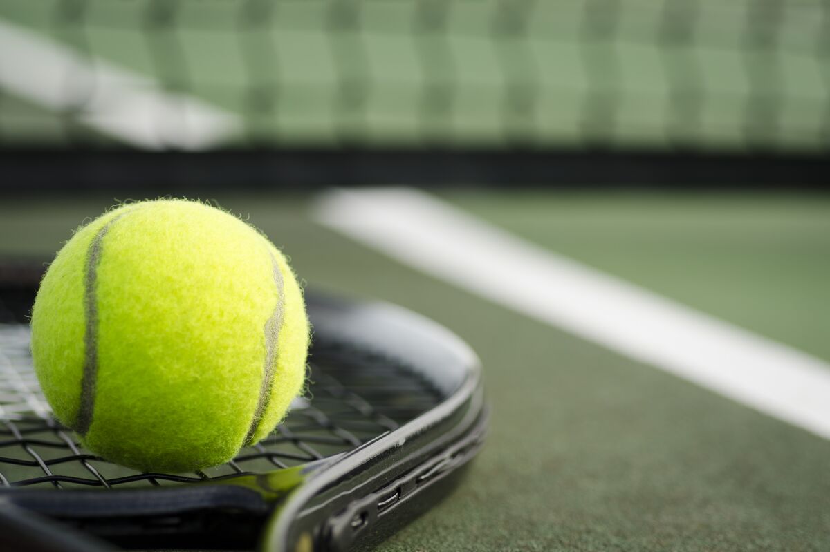 A black tennis racket and yellow tennis ball laying on the ground at a tennis court in early morning light.