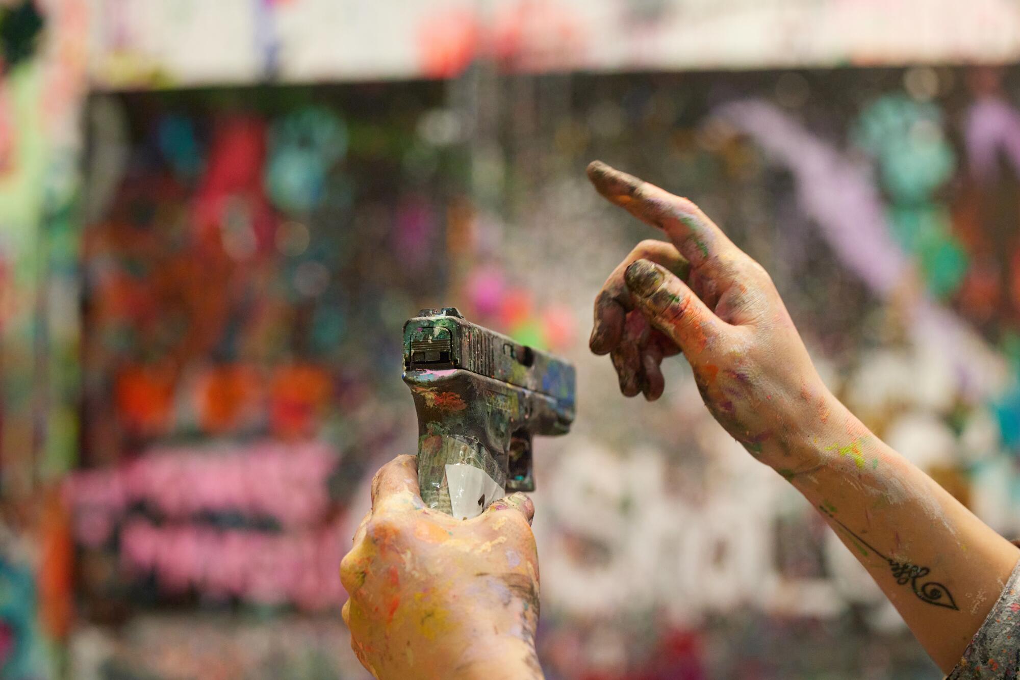 A hand holds a gun covered in multicolor paint.