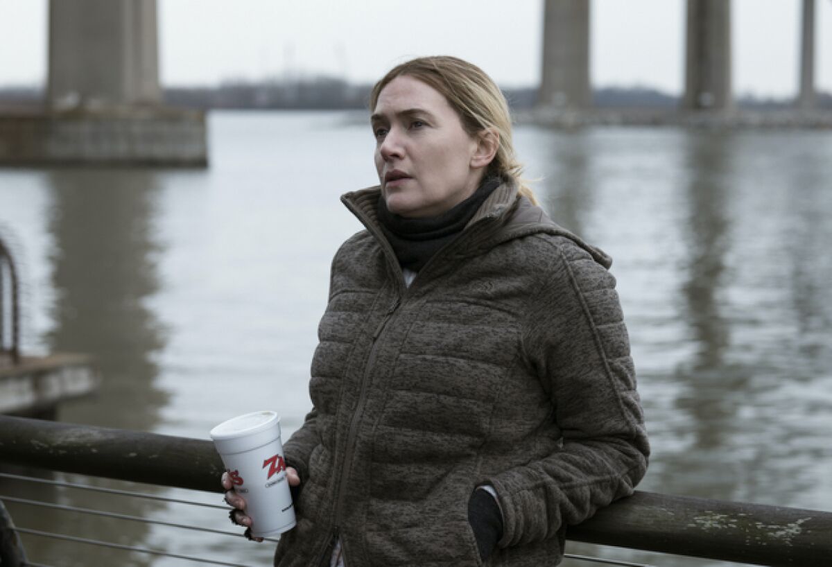 Kate Winslet wears a winter coat and holds a Styrofoam cup beside a river in a scene from "Mare of Easttown."