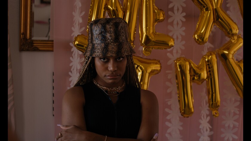 A woman in a bucket hat frowns, gold balloons are in the background