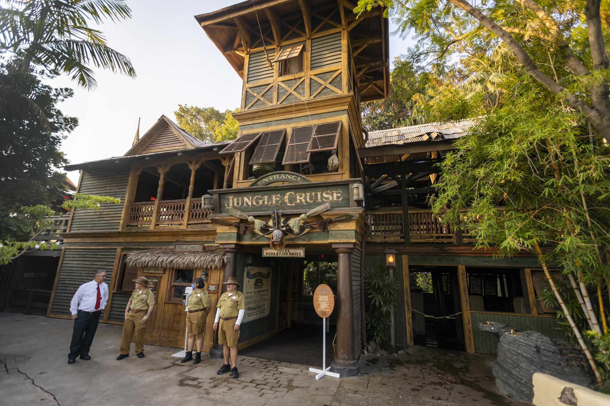 Three people in khaki shorts and shirts and a man in a dress shirt and tie outside a building with a Jungle Cruise sign on it