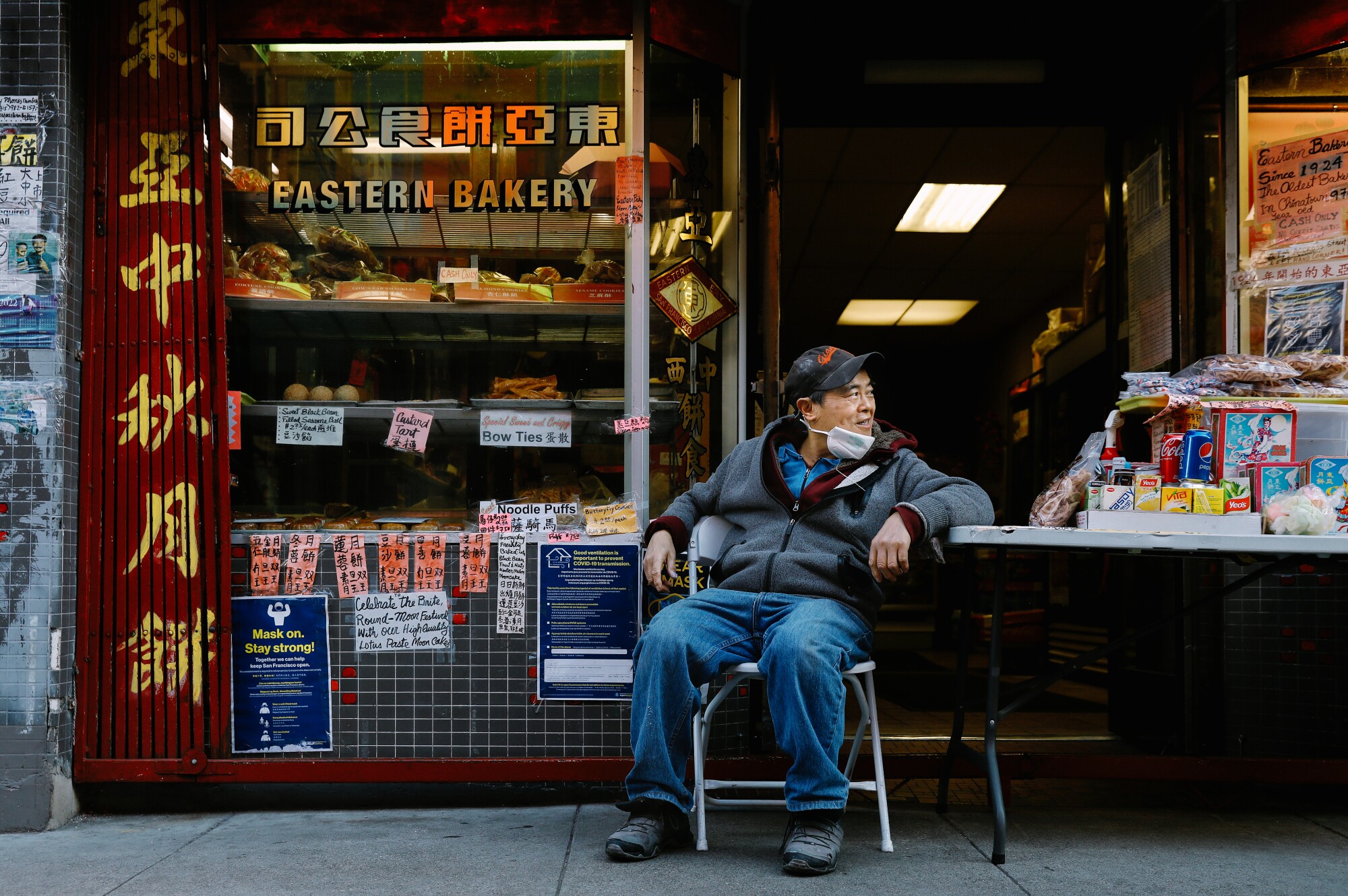 A man sits at a folding table in front of a bakery.