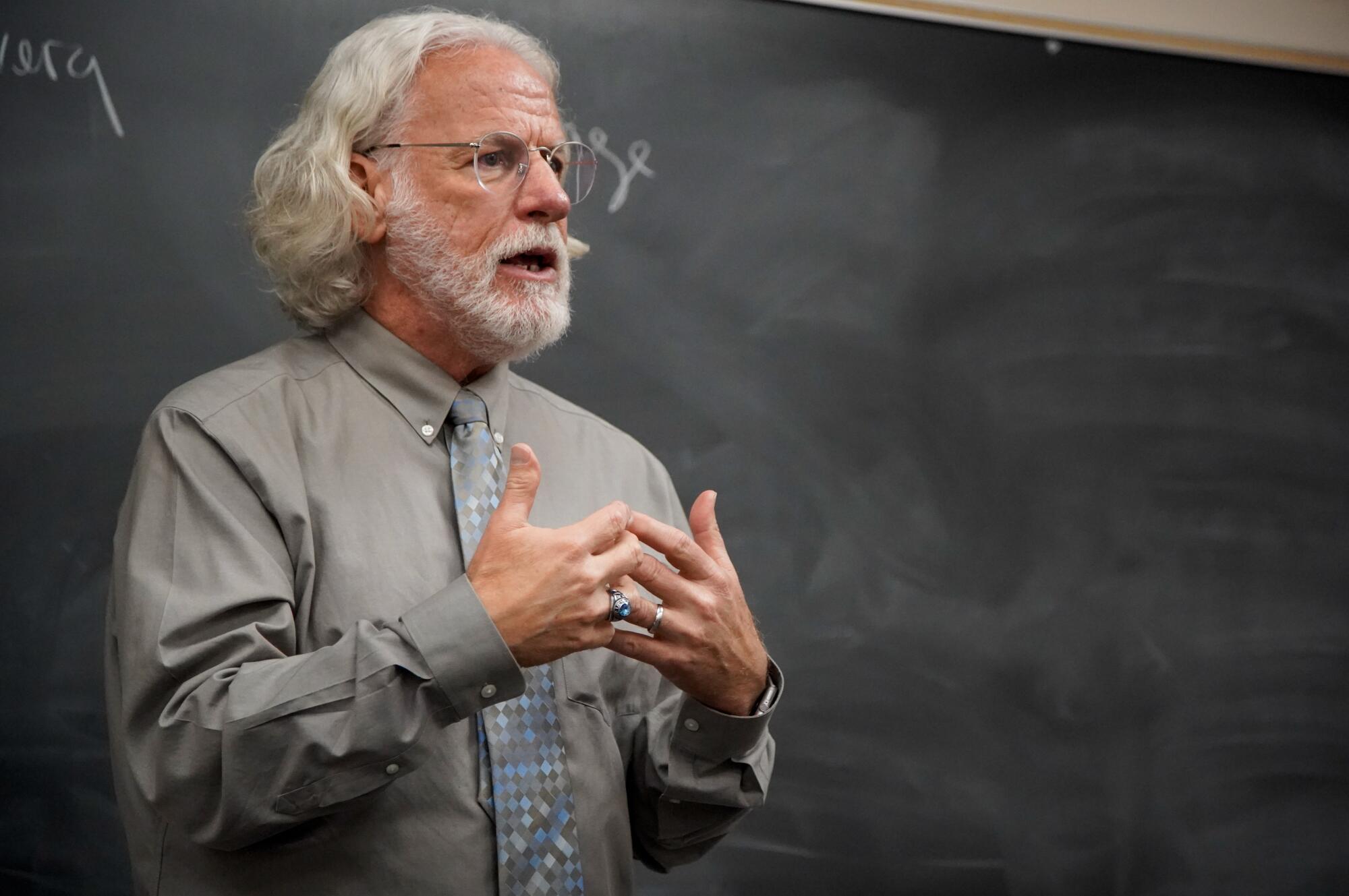 A man with long white hair and beard and wearing glasses stands in front of a chalkboard, gesturing.
