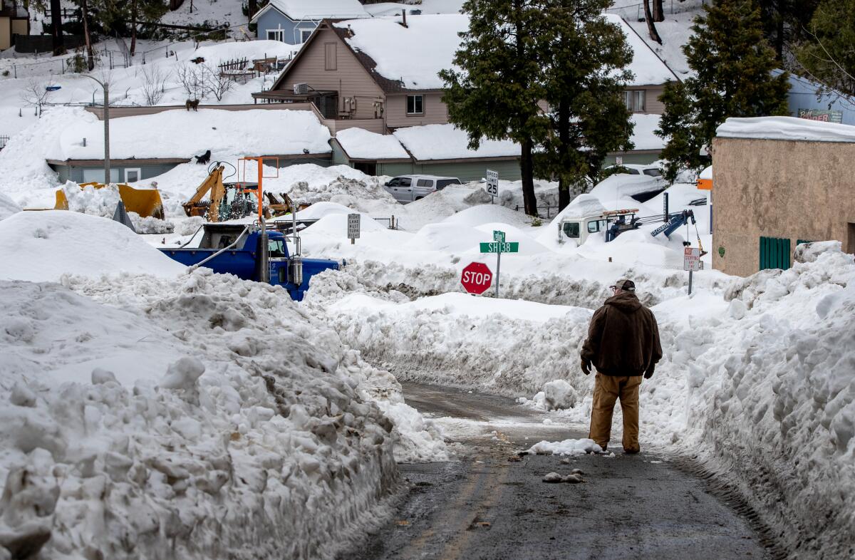 A man stands in a road that has snow stacked up on either side above his head.