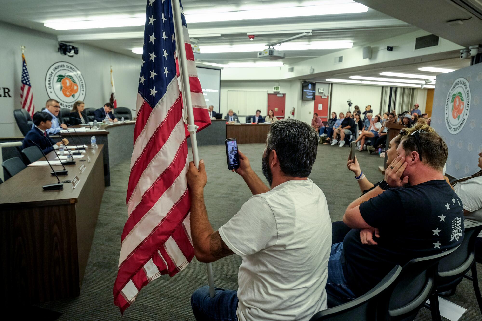 A man holds an American flag as he and another seated nearby hold up cellphones.