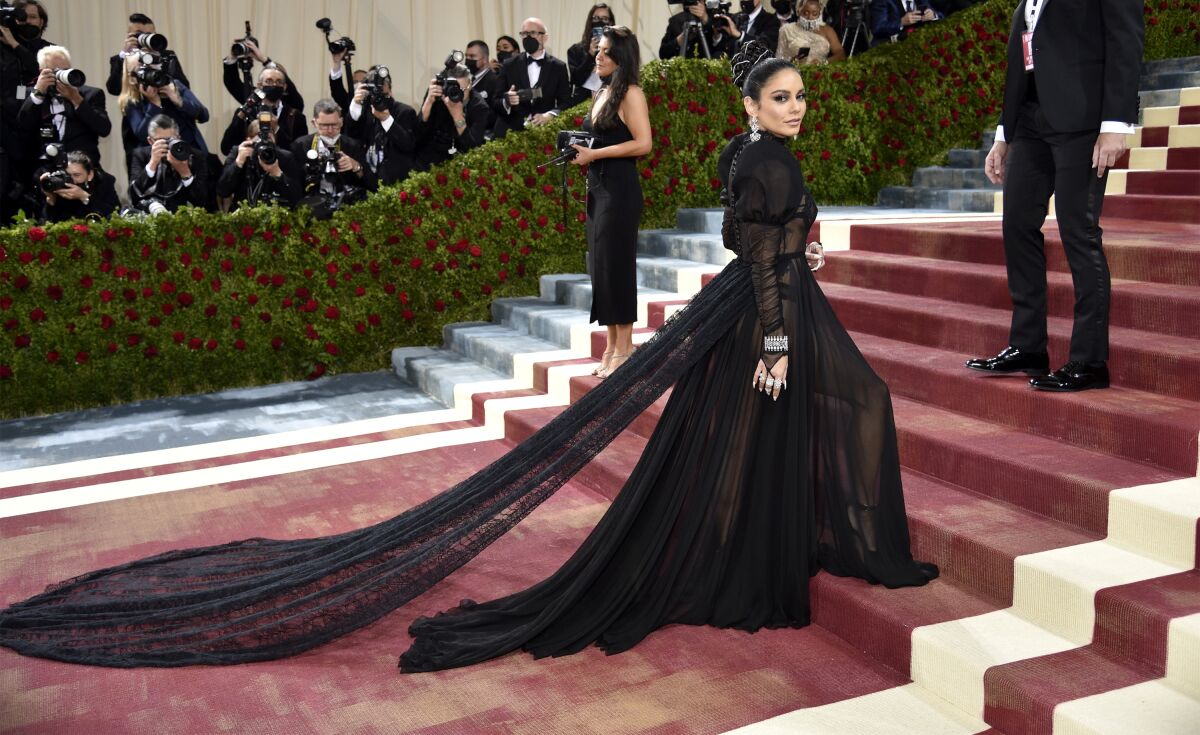 A woman wearing a sheer black gown walks up stairs with photographers in the background.