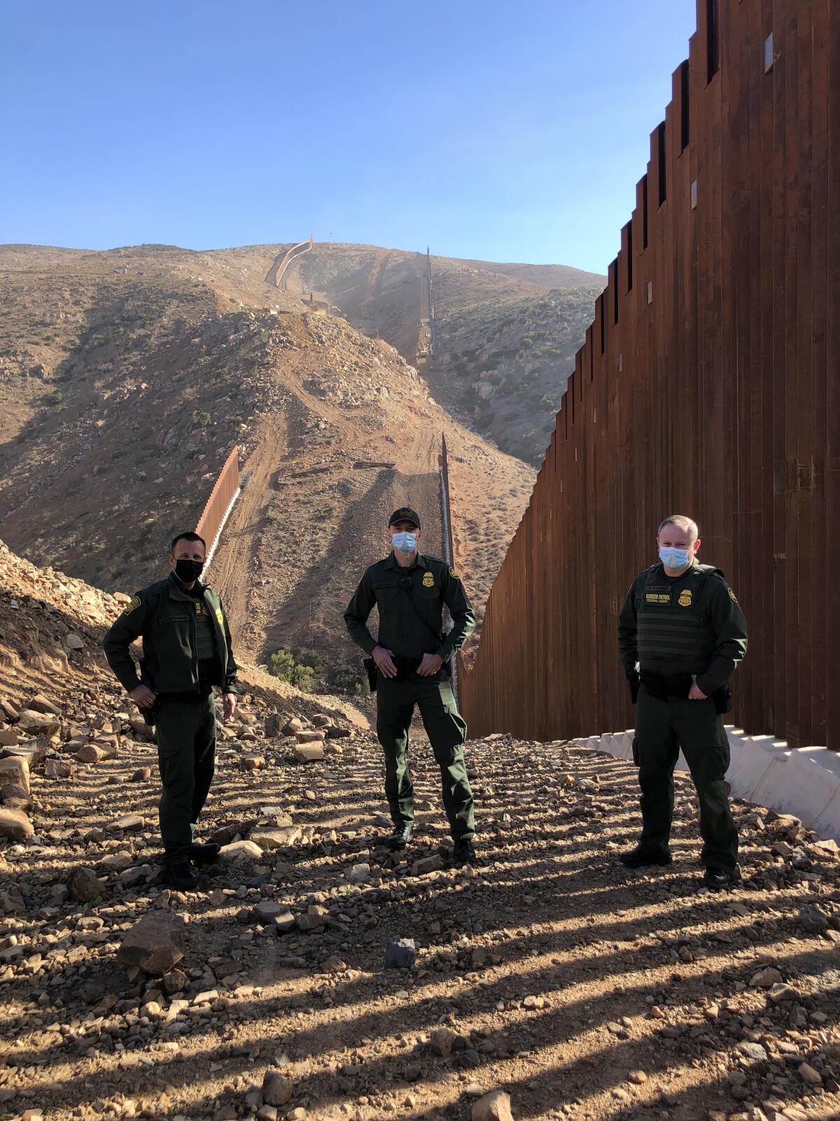 Three border patrol agents stand in rocky dirt next to a border fence in a hilly area