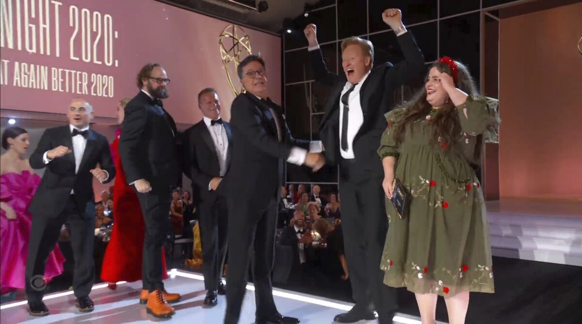 Stephen Colbert turns toward Conan O'Brien, who is raising his arms and smiling, with Aidy Bryant to their right