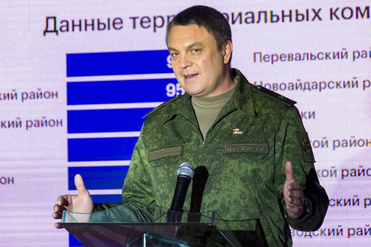 A man in army fatigues with a Z-shaped pin on his chest speaks at a lectern