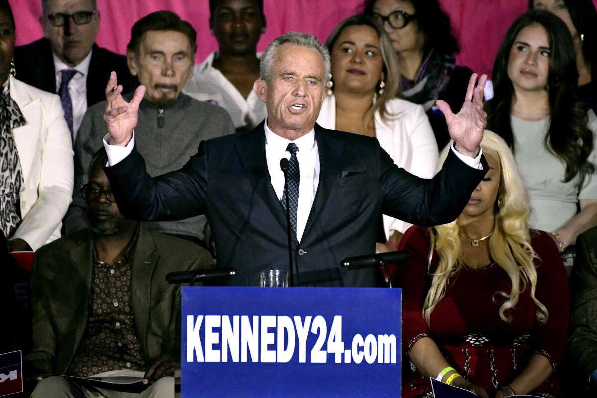 A man with gray hair, dark suit and tie, holds his hands up as he speaks at a lectern 