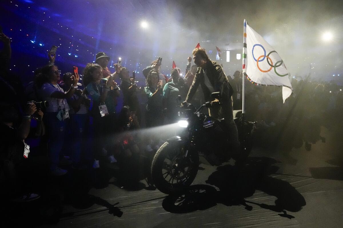 Tom Cruise rides a motorbike with the Olympic flag attached.