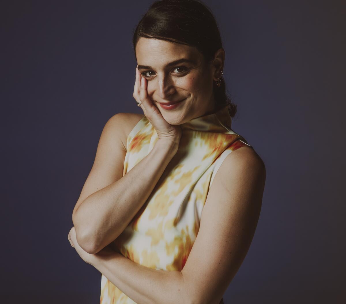 Actor Jenny Slate poses in a sleeveless yellow and white dress, one hand resting on her chin.