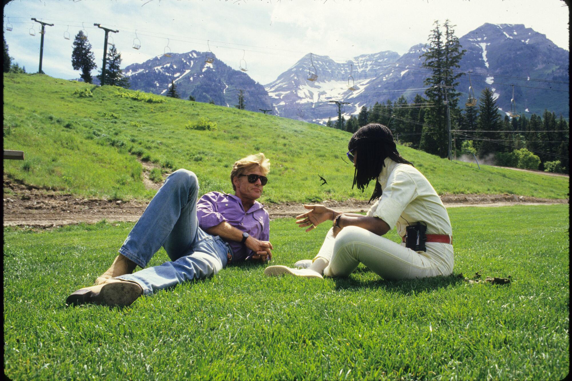 A man, left, and a woman chat in the grass with mountains in the background