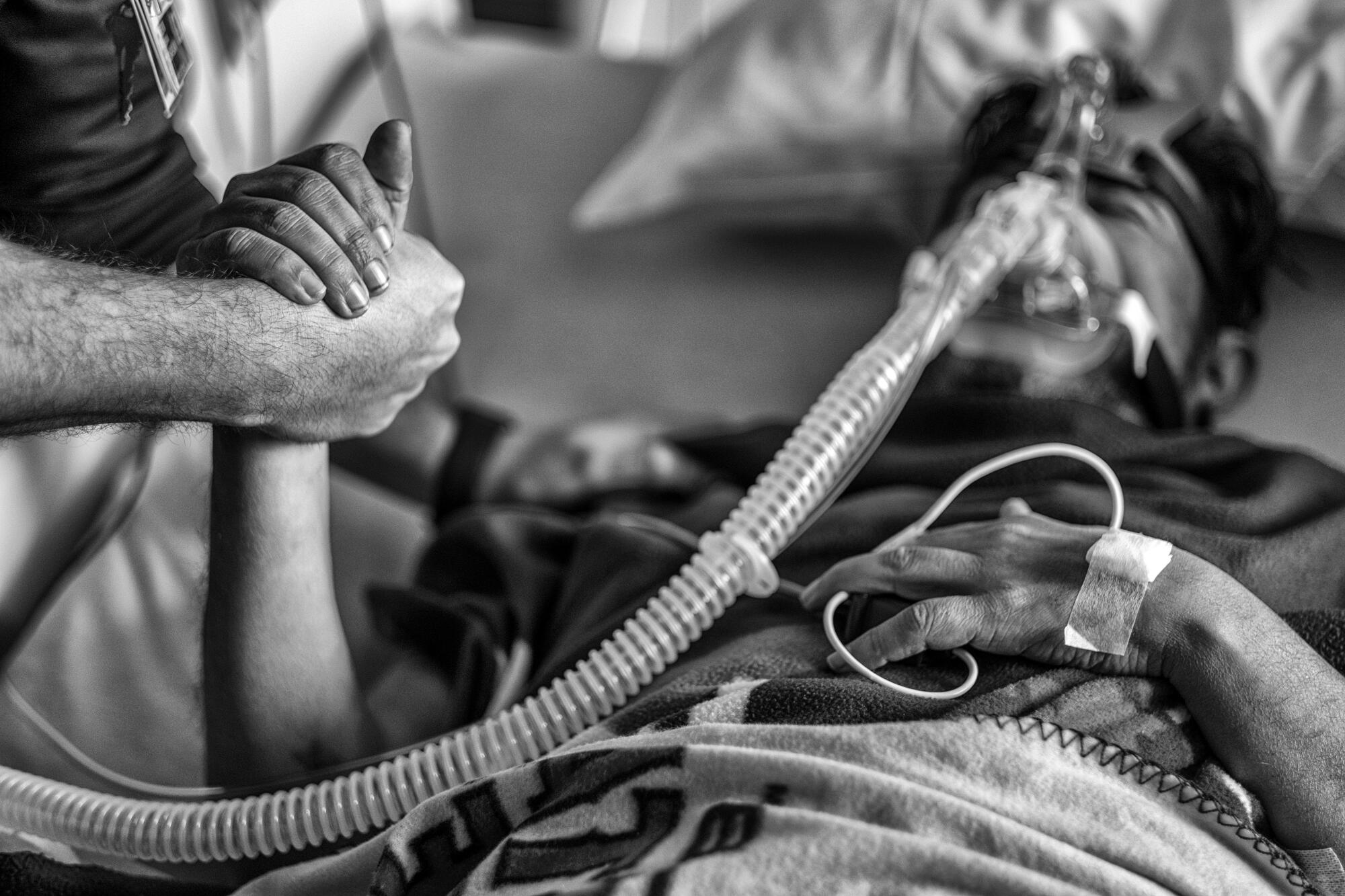 A black-and-white image of a hand reaching in from the left side holding a hand of a patient on respirator in a hospital bed