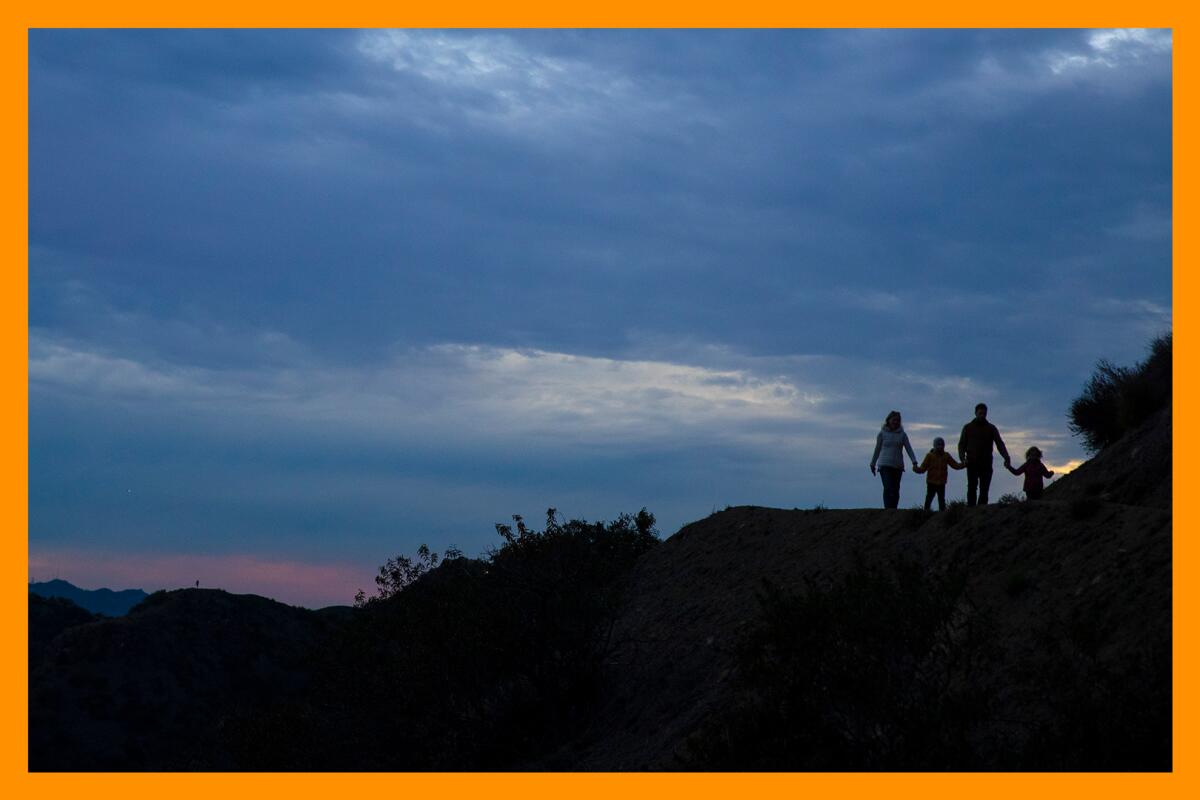 Two adults and two children, all in silhouette on a hiking trail against the backdrop of a setting sun