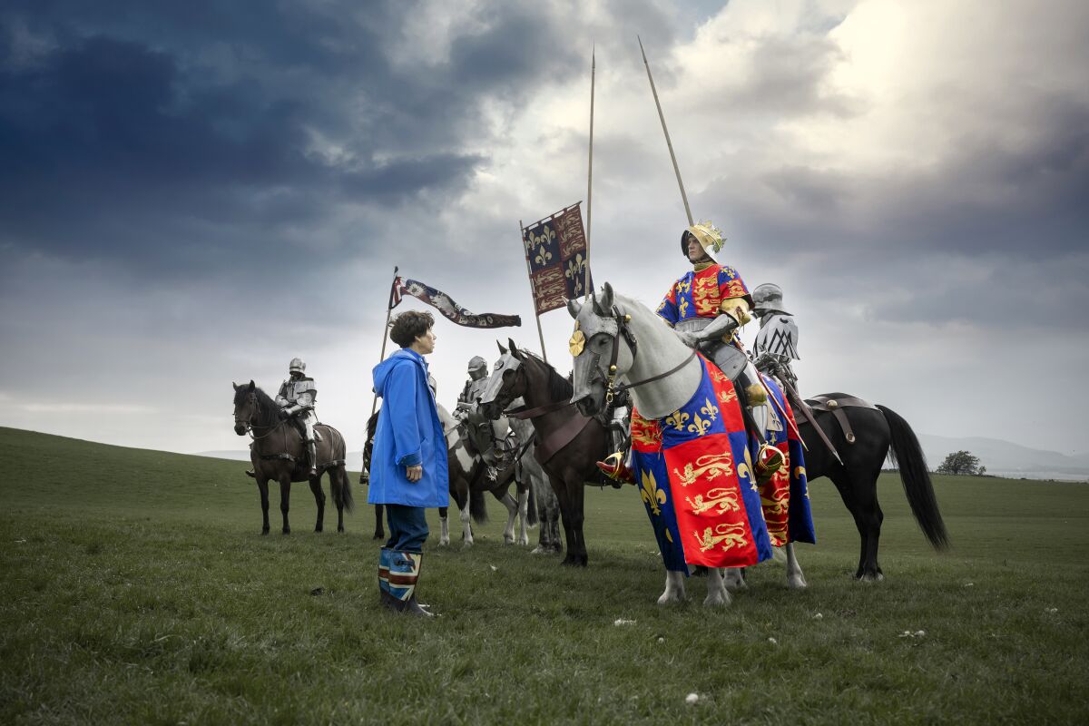 A woman in modern clothing talks with armored horsemen in an open field.