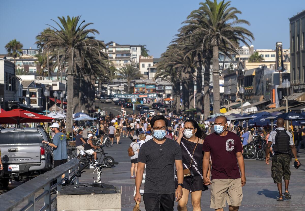 Crowds of people, some in masks, walk along a sidewalk bordered by palm trees.