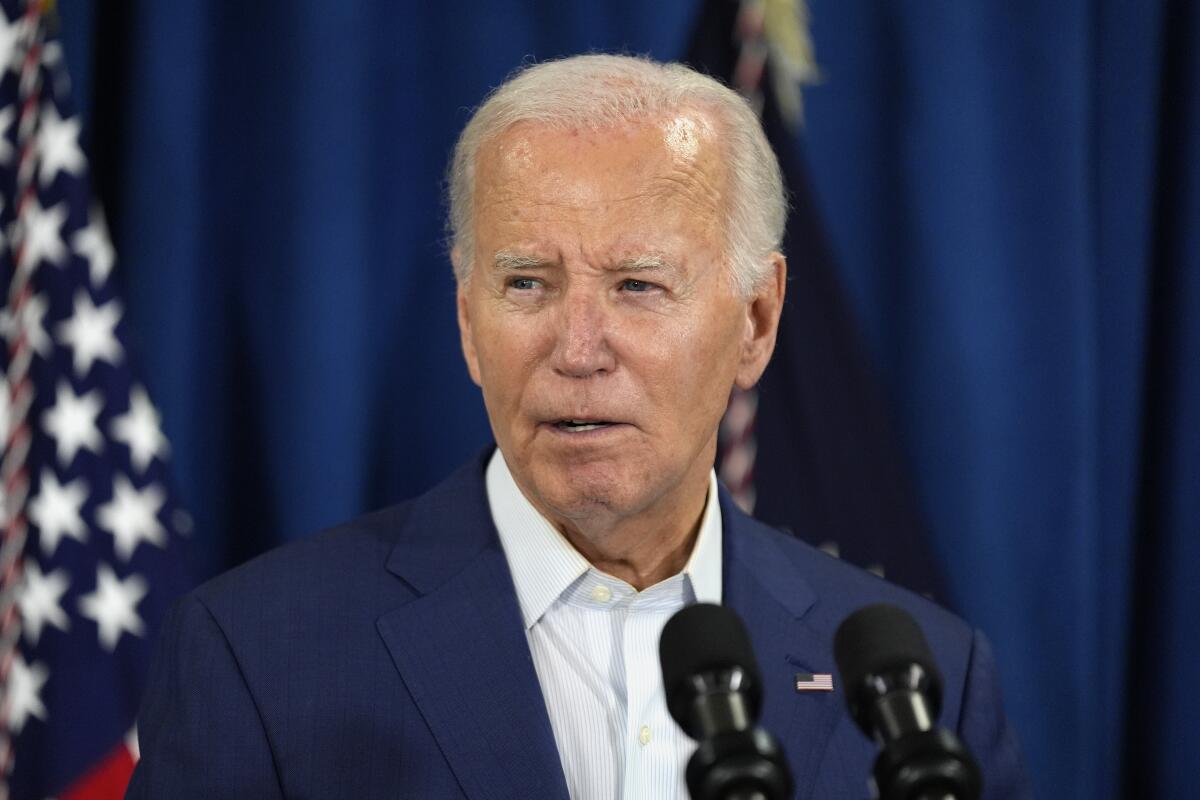 President Biden speaking into two microphones, an American flag in the background