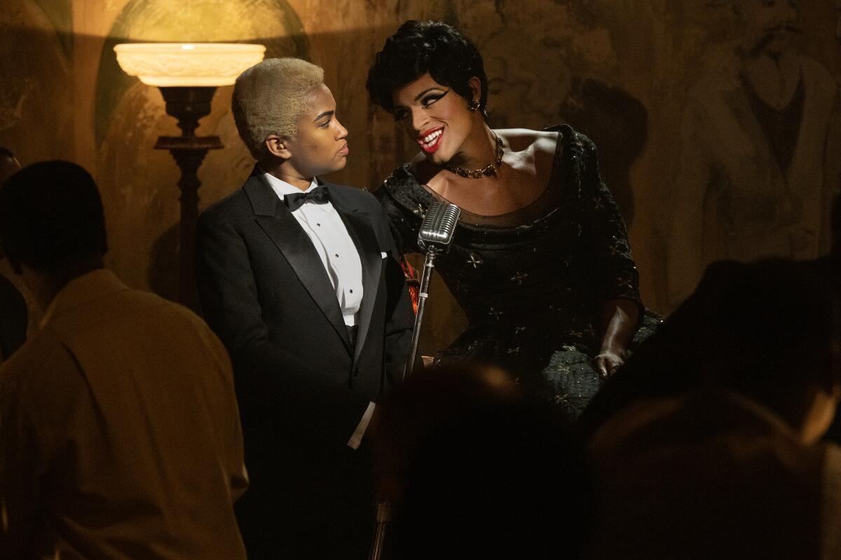 Storme in a black tuxedo stands next to Frankie, who is in drag and singing into a microphone.
