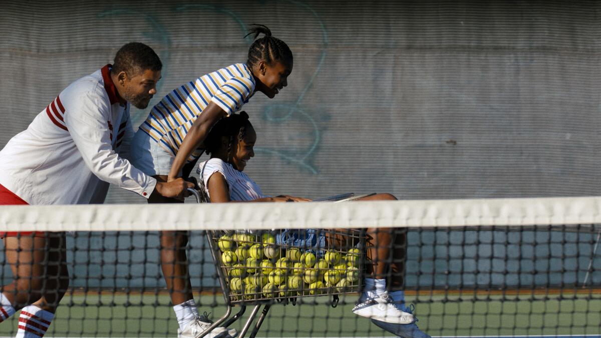 A man pushes a cart of tennis balls with two girls on it in a scene in 'King Richard.'