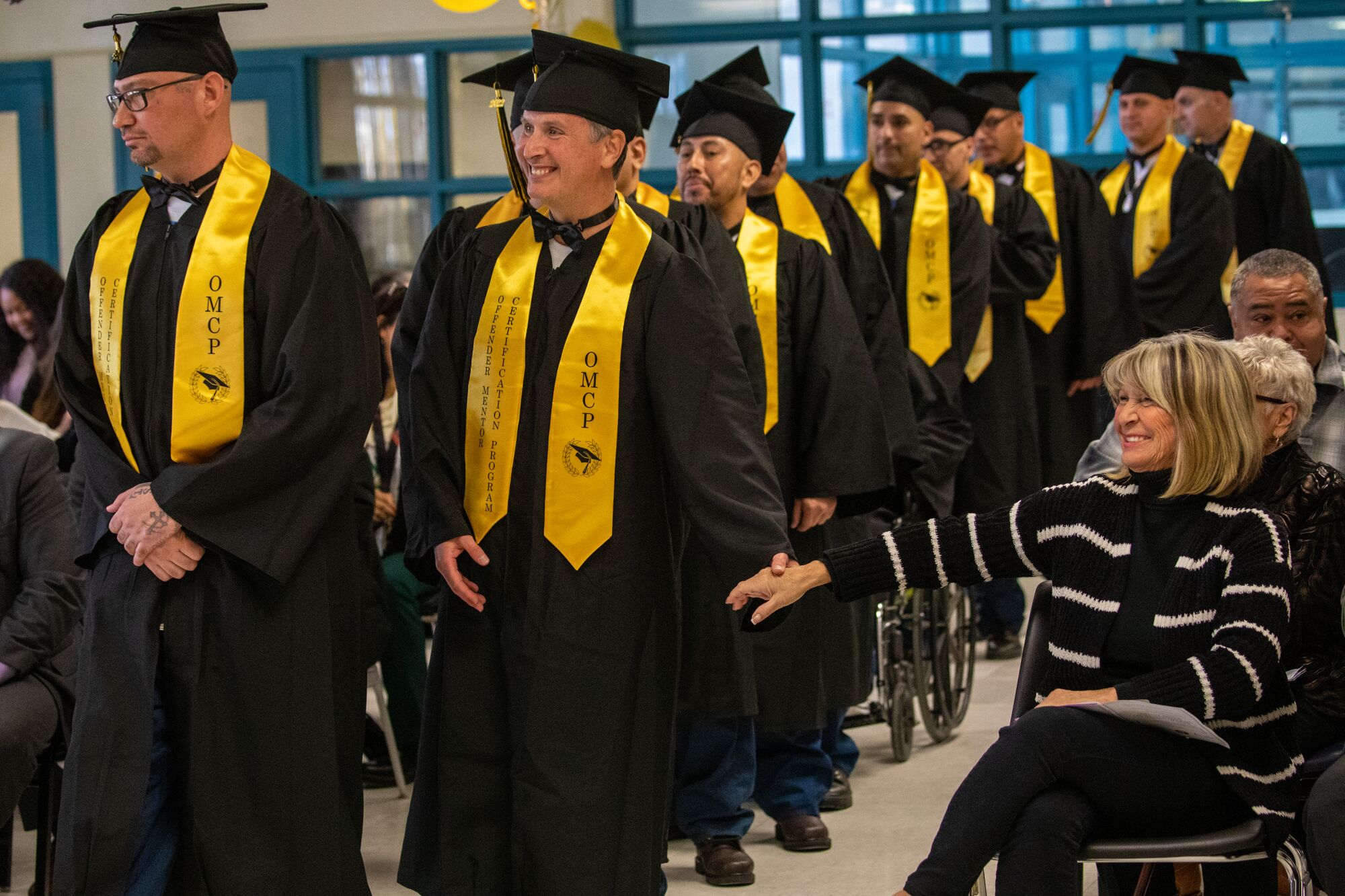 men in caps and gowns and yellow stoles smile as they walk down an aisle