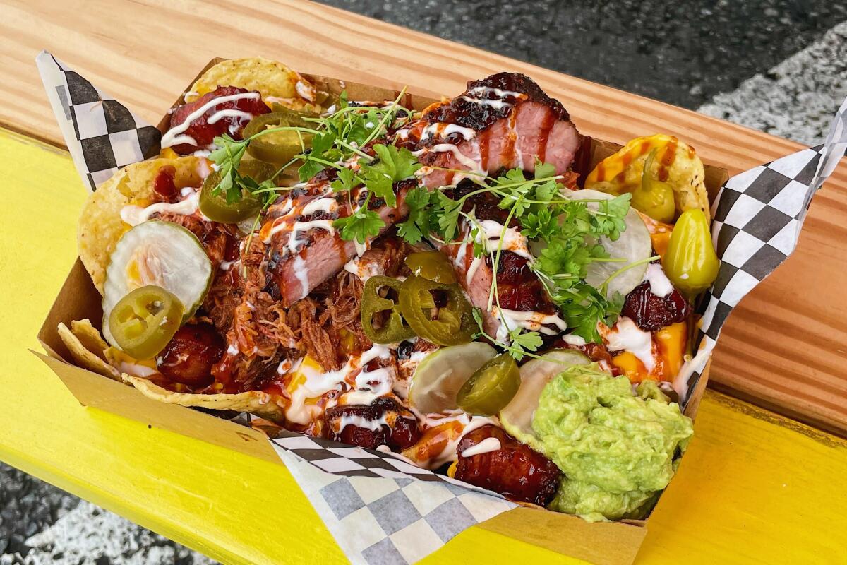 A to-go box of  Battambong BBQ nachos piled with pulled pork, pickles, ribs and pork belly on a yellow wooden bench