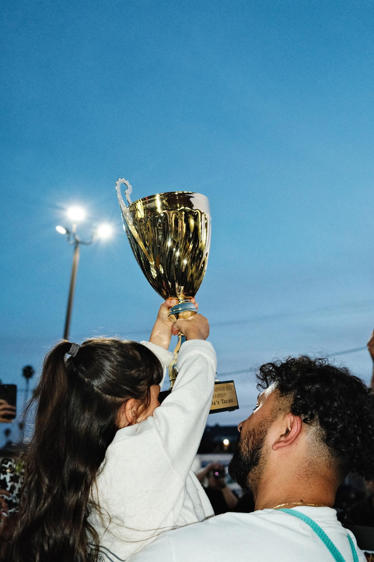 A little girl, seen from behind, holds a large gold trophy overhead in a parking lot at sundown.