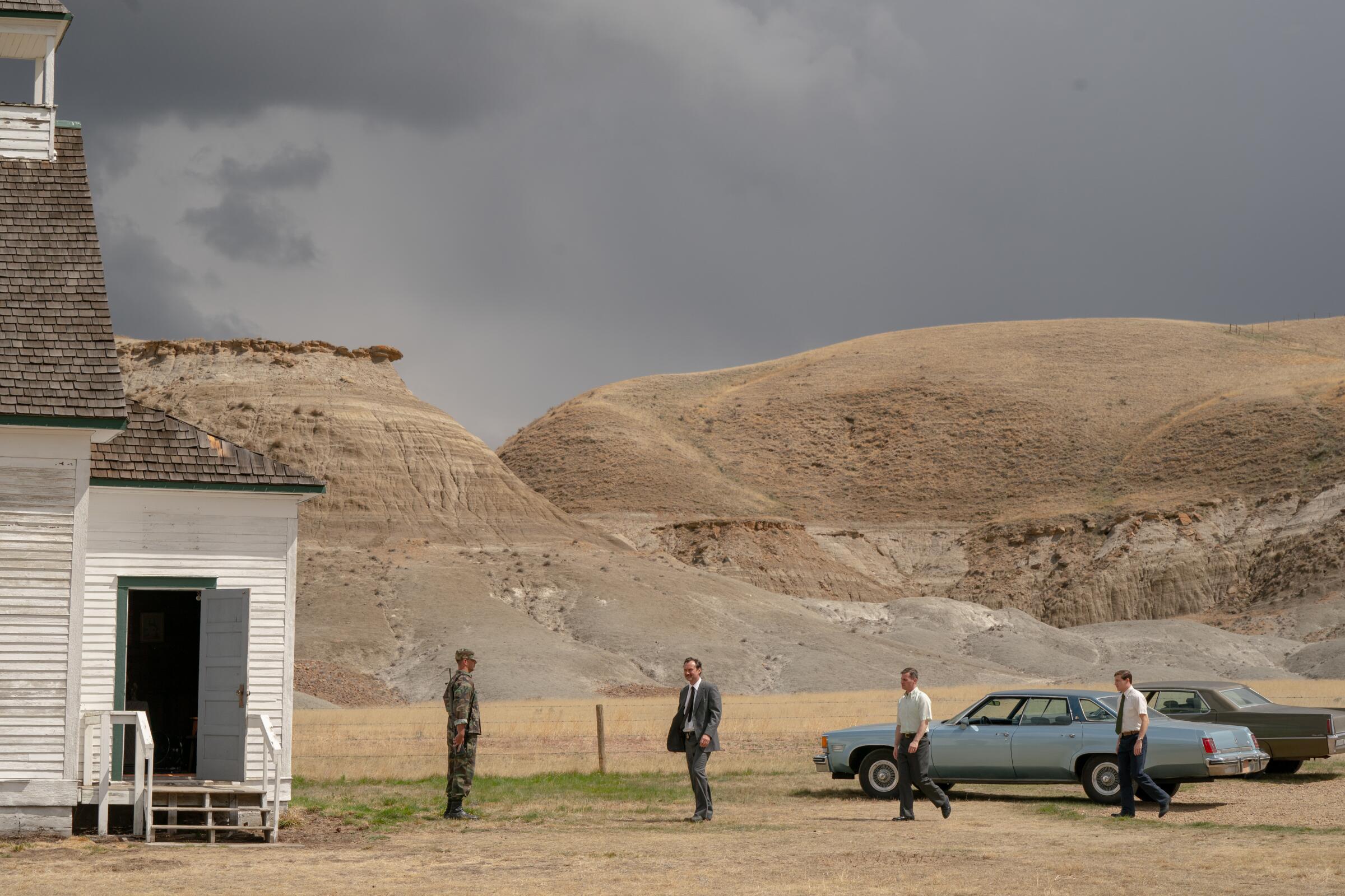 A group of men stand between two parked cars and a white clapboard house in the wilderness.