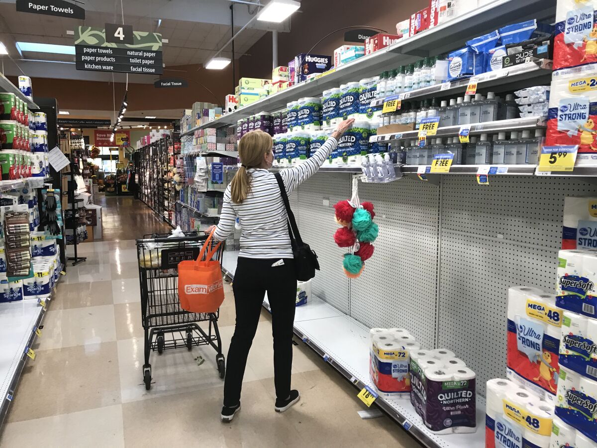 A woman pushing a cart in a grocery aisle reaches for toilet paper amid partially emptied shelves.