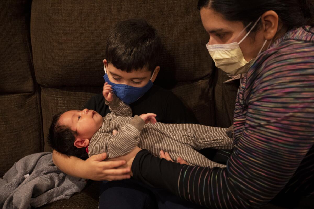 A woman in a mask puts a newborn boy in the arms of a toddler, also wearing a mask