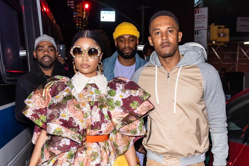 Nicki Minaj in large, round sunglasses and a bulky floral print dress standing next to a man in a beige hoodie and pants