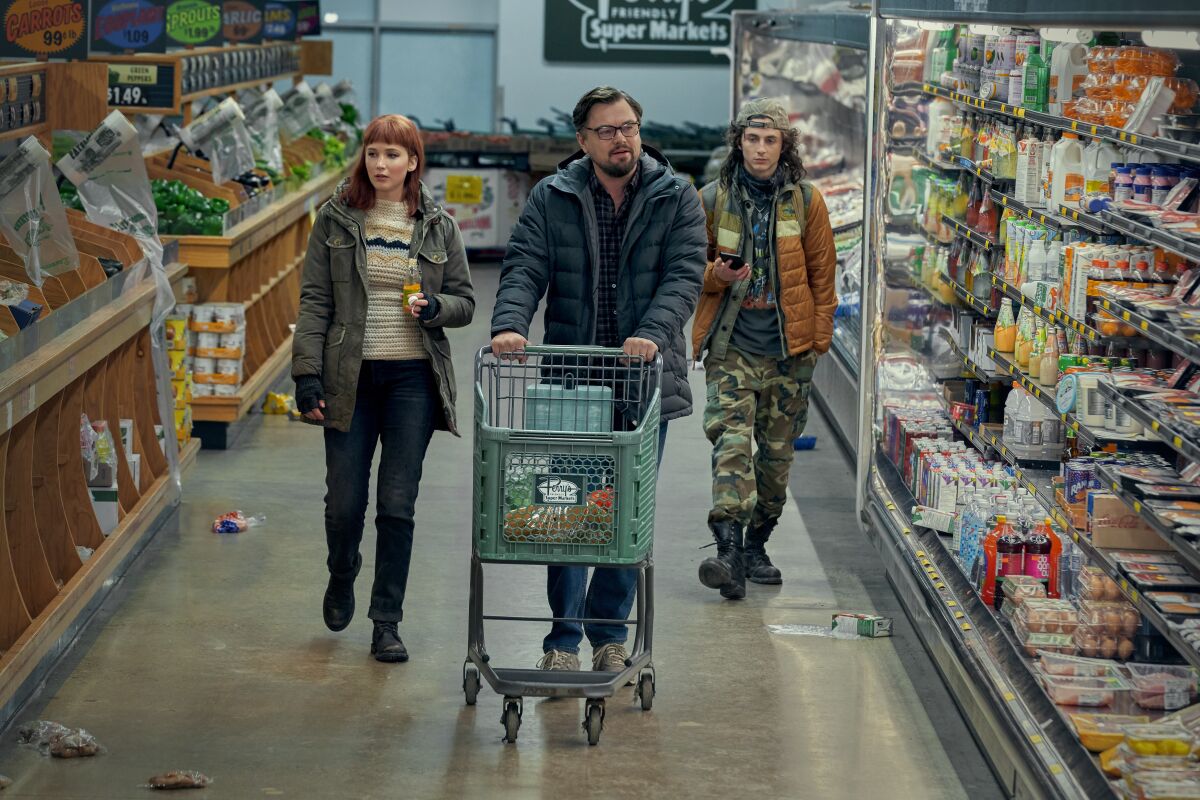 A man pushes a cart through a grocery store, followed by a man and a woman.