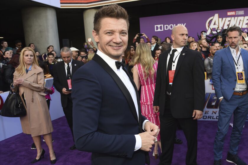 A man with short brown hair in a black tuxedo smiling among a crowd of people on a purple carpet