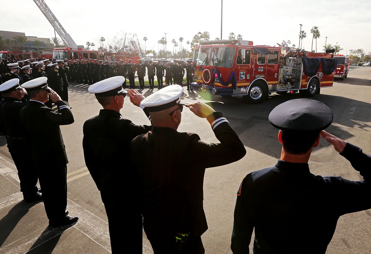 Rows of firefighters in dress uniforms salute a passing firetruck