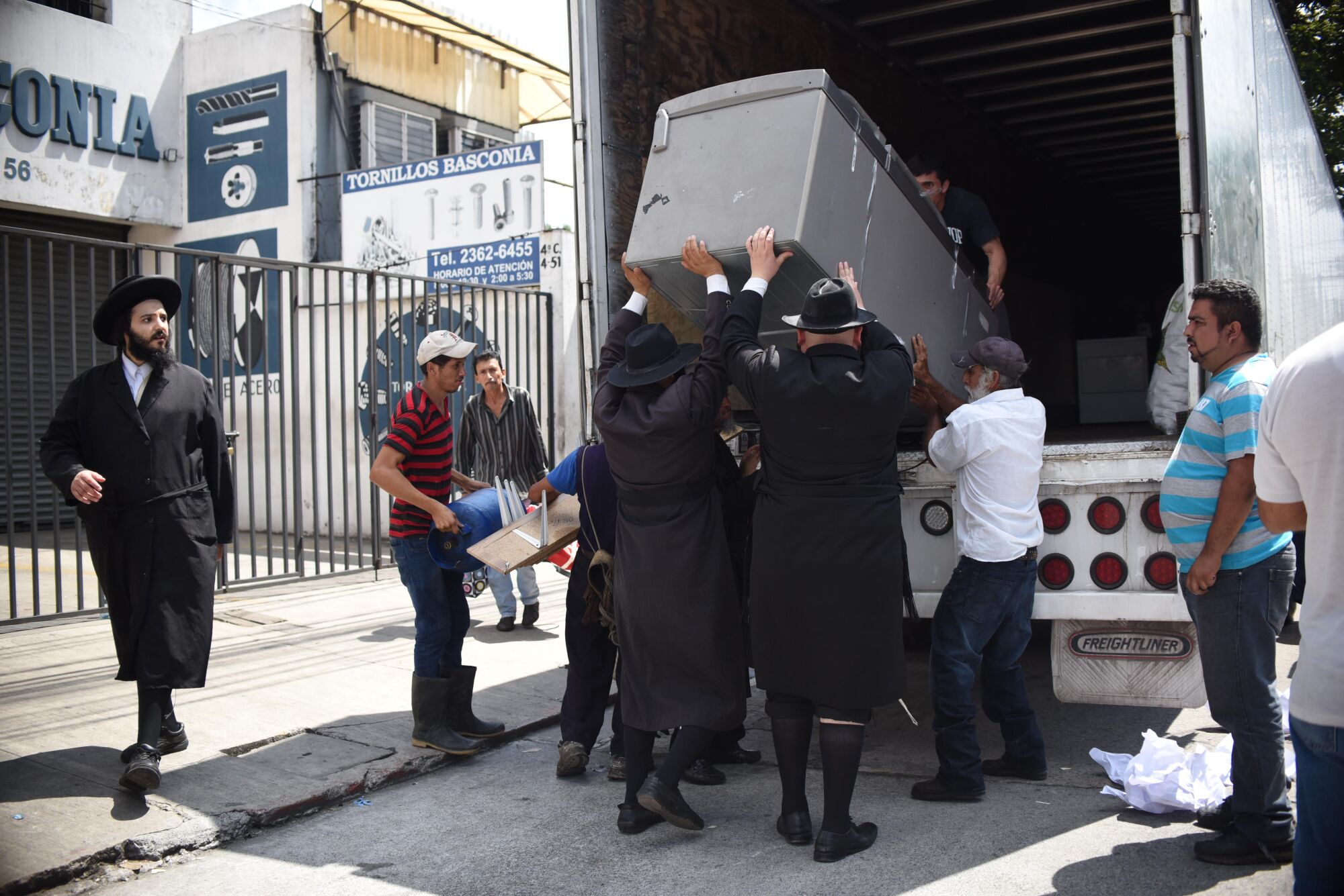 A bearded man, left, in dark clothes and hat looks at a group of men hoisting a long gray container at the back of a truck 