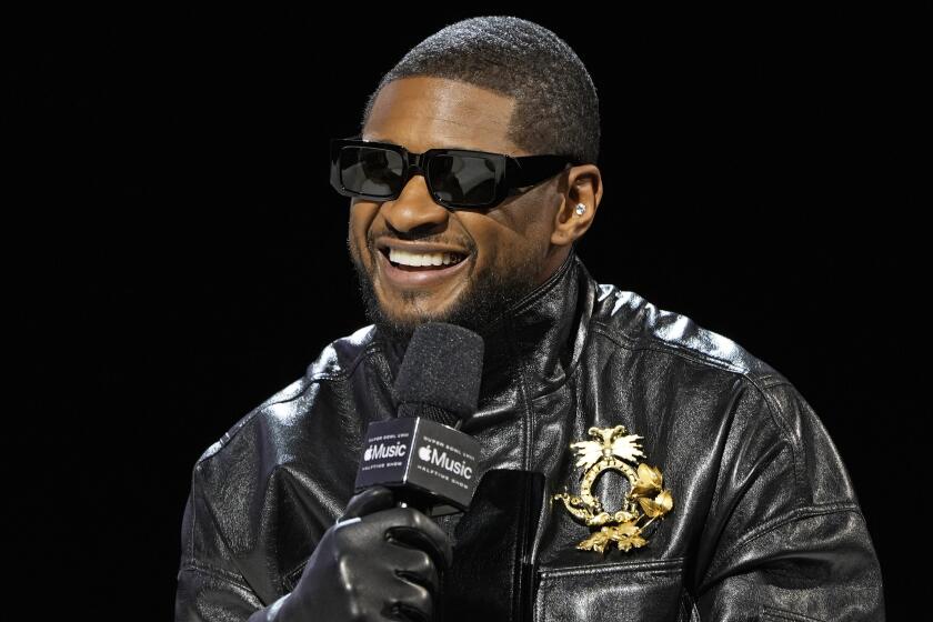 Usher smiling and wearing big square sunglasses, a black leather jacket and gloves. He is holding a microphone 
